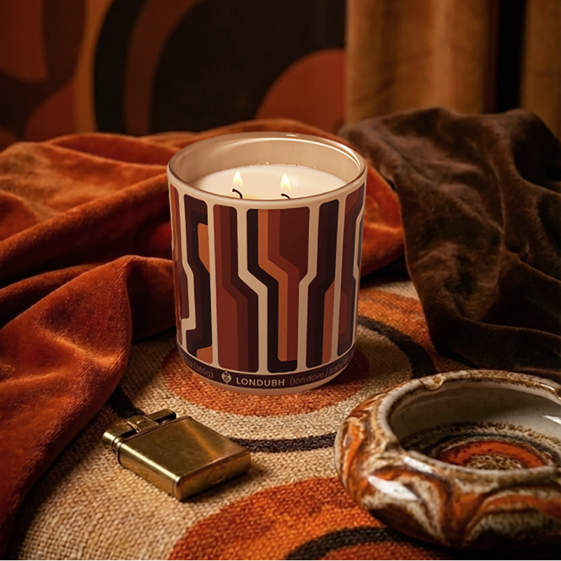 The lit Disco Biscuit Candle in a retro scene with an orange shag throw, a gold lighter, and a ceramic ashtray.