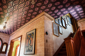 Peacock Nouveau aubergine purple, pink, and gold wallpaper shown installed on ceiling of house foyer and staircase against stone walls. Peacock Nouveau Aubergine is a Celtic and Art Nouveau inspired maximalist repeat pattern wallpaper featuring peacocks and Celtic knot work in pinks, purples, all outlined in gold and digitally printed on gold mylar.