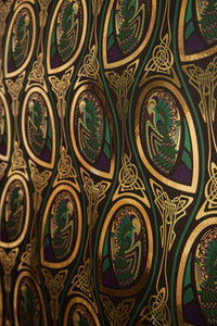 Peacock Nouveau Bejeweled is a Celtic and Art Nouveau inspired maximalist repeat pattern wallpaper featuring peacocks and Celtic knot work in gold, jewel tones, blues, greens, on a black background, outlined in gold and digitally printed on gold mylar - shown at an angle with the light bouncing off the gold