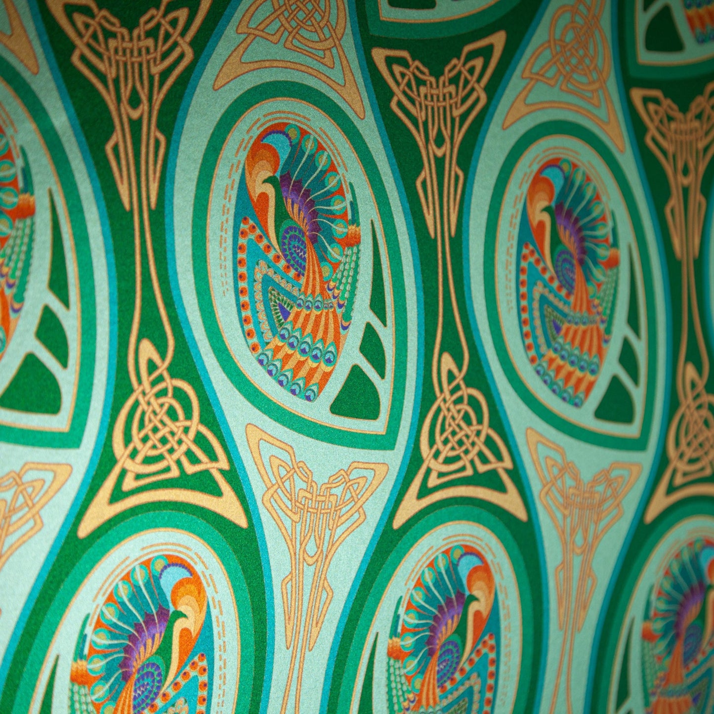 Peacock Nouveau Green Garden is a shimmering Celtic and Art Nouveau inspired maximalist repeat pattern wallpaper featuring peacocks and Celtic knot work in various greens, orange, blue, yellow, blues, purple, on a light green background, digitally printed on silver mylar - shown at an angle to highlight the shimmer