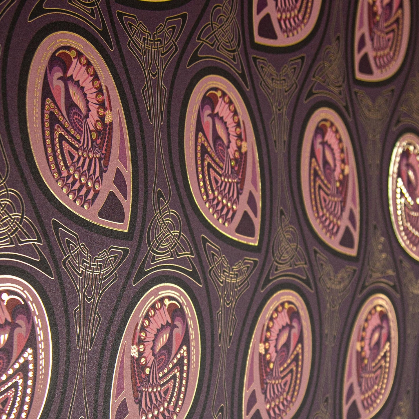 Peacock Nouveau Aubergine is a Celtic and Art Nouveau inspired maximalist repeat pattern wallpaper featuring peacocks and Celtic knot work in pinks, purples, all outlined in gold and digitally printed on gold mylar - shown at an angle with the light catching off the gold