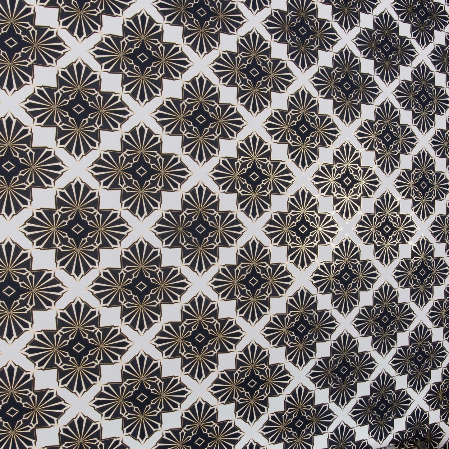 An angle shot of Blaze Inhale, a repeating geometric pattern featuring four black and white cannabis and marijuana leaves thinly framed in copper brown on a white background digitally printed on gold mylar shown illuminated in light bouncing off the shimmering gold trim outlining the pattern