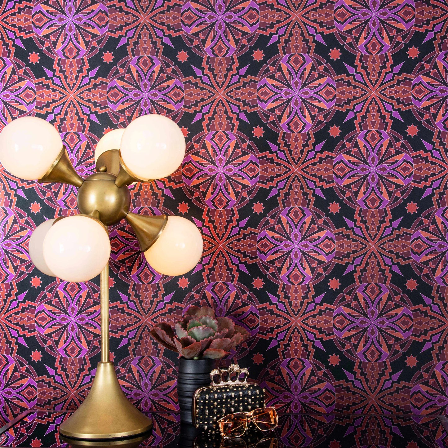 ChaCha Chill is a 1920s art deco inspired shimmering repeat geometric pattern composed of stars, diamonds, triangles, and other shapes in 70s retro pinks, reds, oranges, burgundy, on a black background and digitally printed on silver mylar - shown in a vignette with a deco lamp, a plant, vintage Ray Ban sunglasses and a black leather Alexander McQueen clutch bedazzled in skulls.