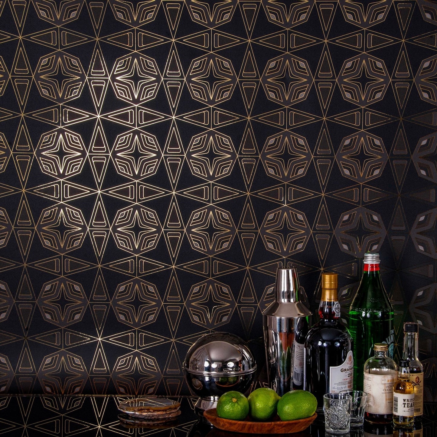 Integratronic Night is a mid-century retro repeat geometric pattern featuring dark green and gray motifs on a black background all outlined in gold and digitally printed on gold mylar - shown in a vignette with a bar set up with bottles, limes, and shot glasses