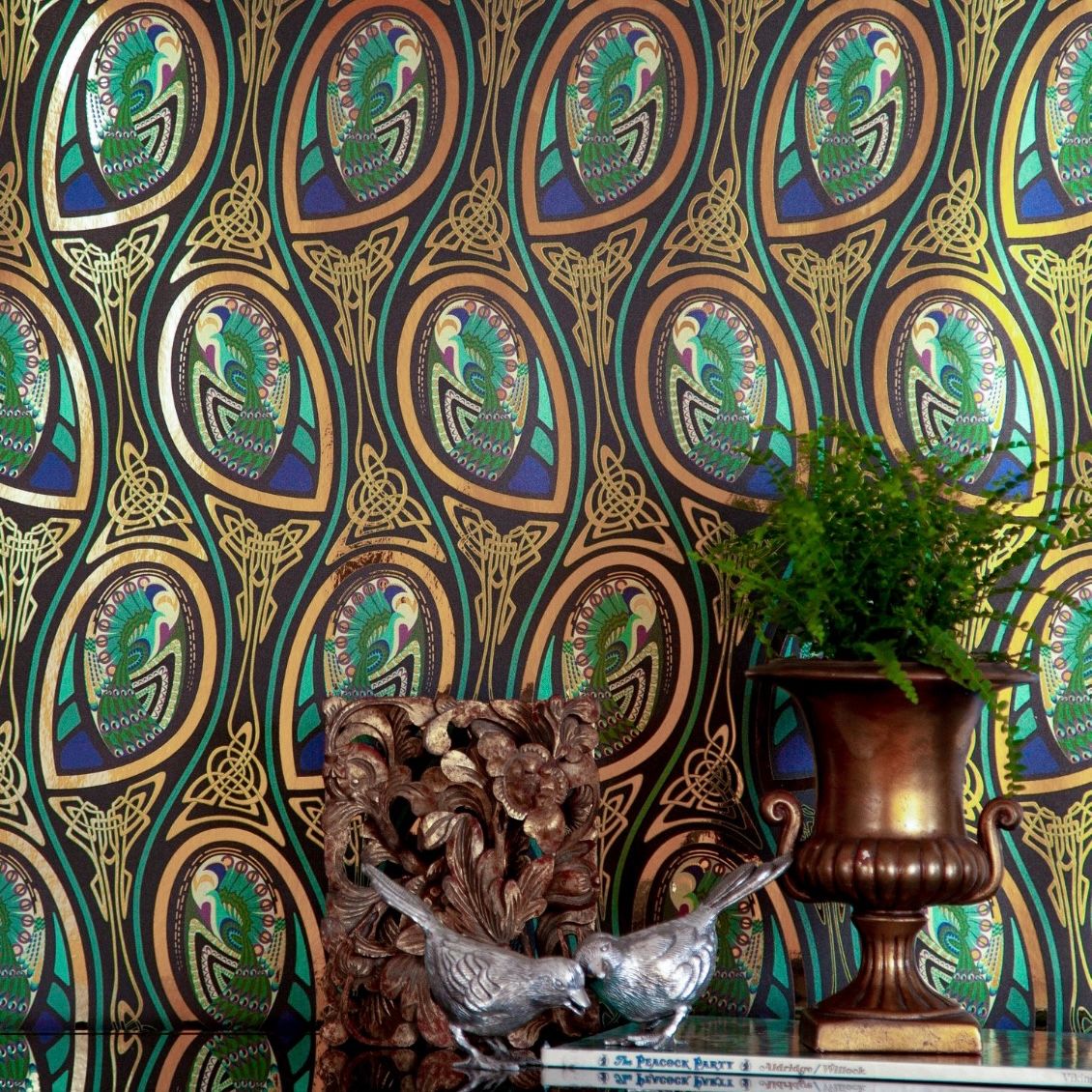 Peacock Nouveau Bejeweled is a Celtic and Art Nouveau inspired maximalist repeat pattern wallpaper featuring peacocks and Celtic knot work in gold, jewel tones, blues, greens, on a black background, outlined in gold and digitally printed on gold mylar  - shown in a vignette with plants, a block of gnarled wood, a book and a metal sculpture of 2 birds