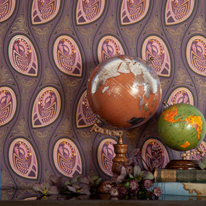 Peacock Nouveau Aubergine is a Celtic and Art Nouveau inspired maximalist repeat pattern wallpaper featuring peacocks and Celtic knot work in pinks, purples, all outlined in gold and digitally printed on gold mylar - shown in a vignette with vintage books and globes