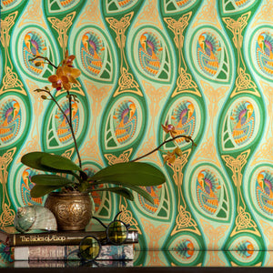 Peacock Nouveau Green Garden is a shimmering Celtic and Art Nouveau inspired maximalist repeat pattern wallpaper featuring peacocks and Celtic knot work in various greens, orange, blue, yellow, blues, purple, on a light green background, digitally printed on silver mylar - shown in a vignette with vintage books, an orchid, and a shell