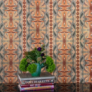  Sampa Terra is a riff of a classical vintage repeat linear geometric pattern wallpaper in earth tones, greens and oranges, with all shapes outlined in gold  digitally printed on gold mylar - shown in a vignette with flowers and coffee table books.