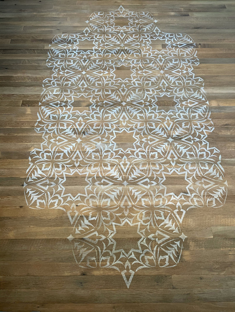 A large, intricate geometric stencil in white creates a runner-like pattern on a light wood floor.