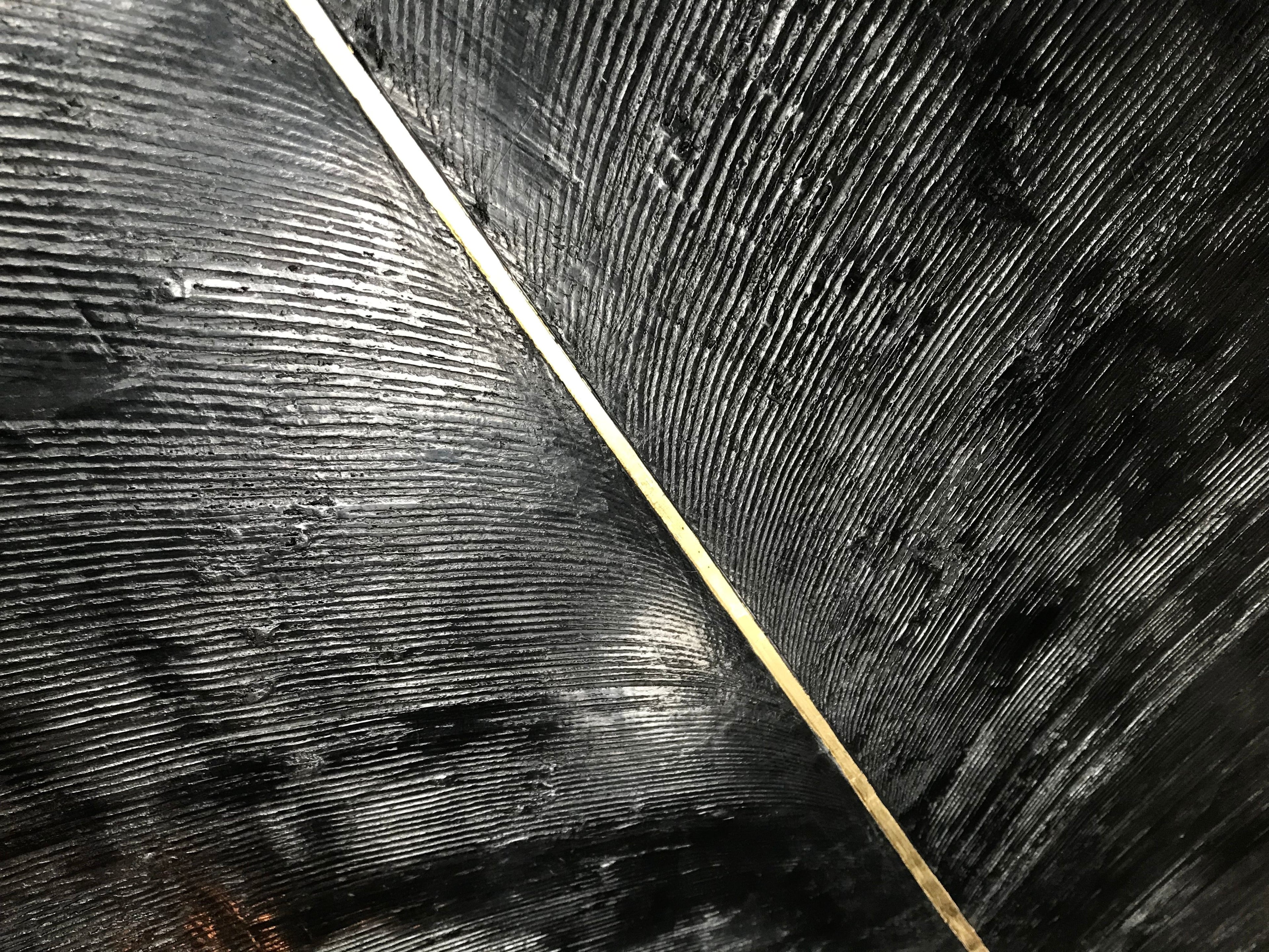 A close-up of a textured black art piece with deep, curved grooves bisected by a thin diagonal gold line.