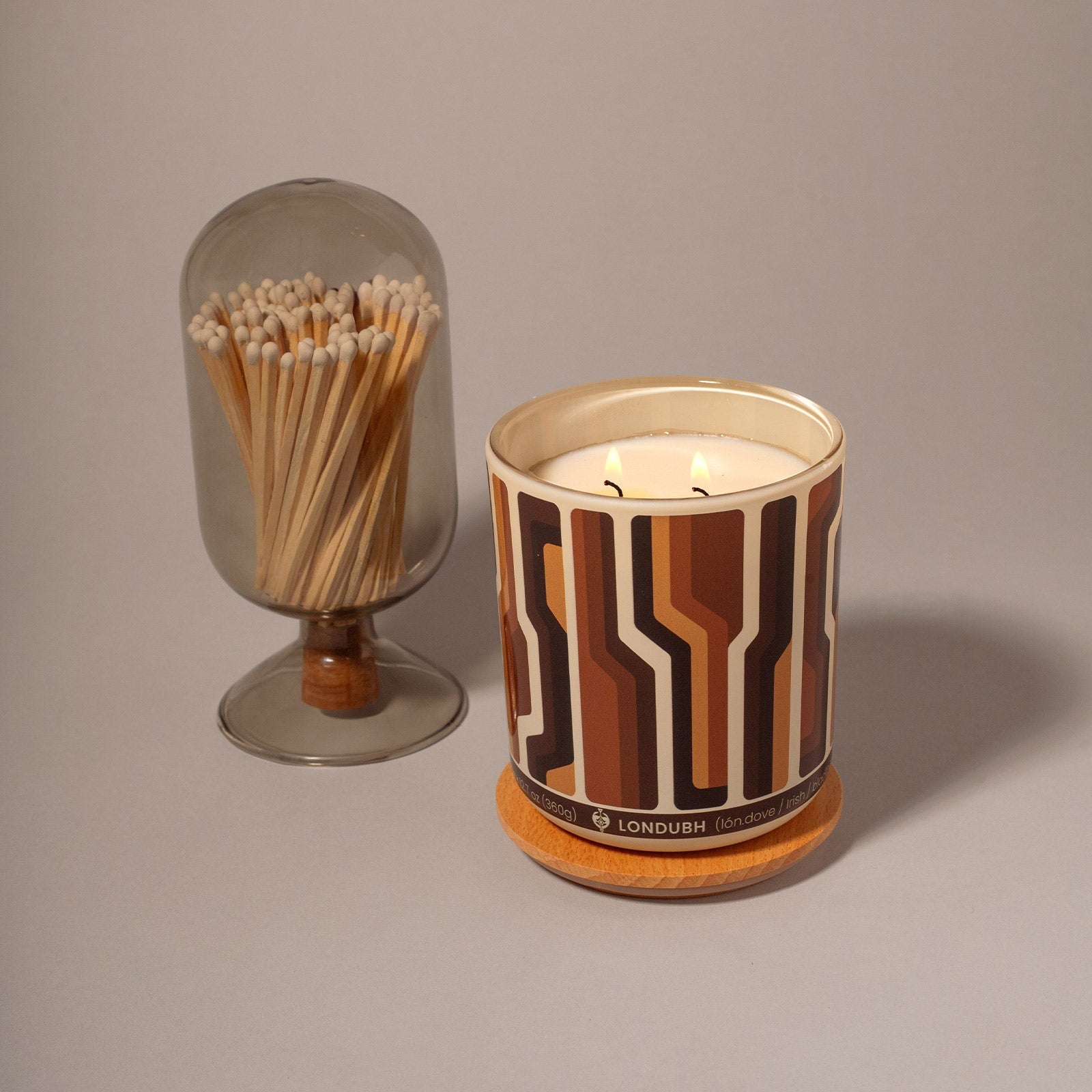 The lit Disco Biscuit Candle sits on a cork coaster next to a smoky glass cloche filled with wooden matches.