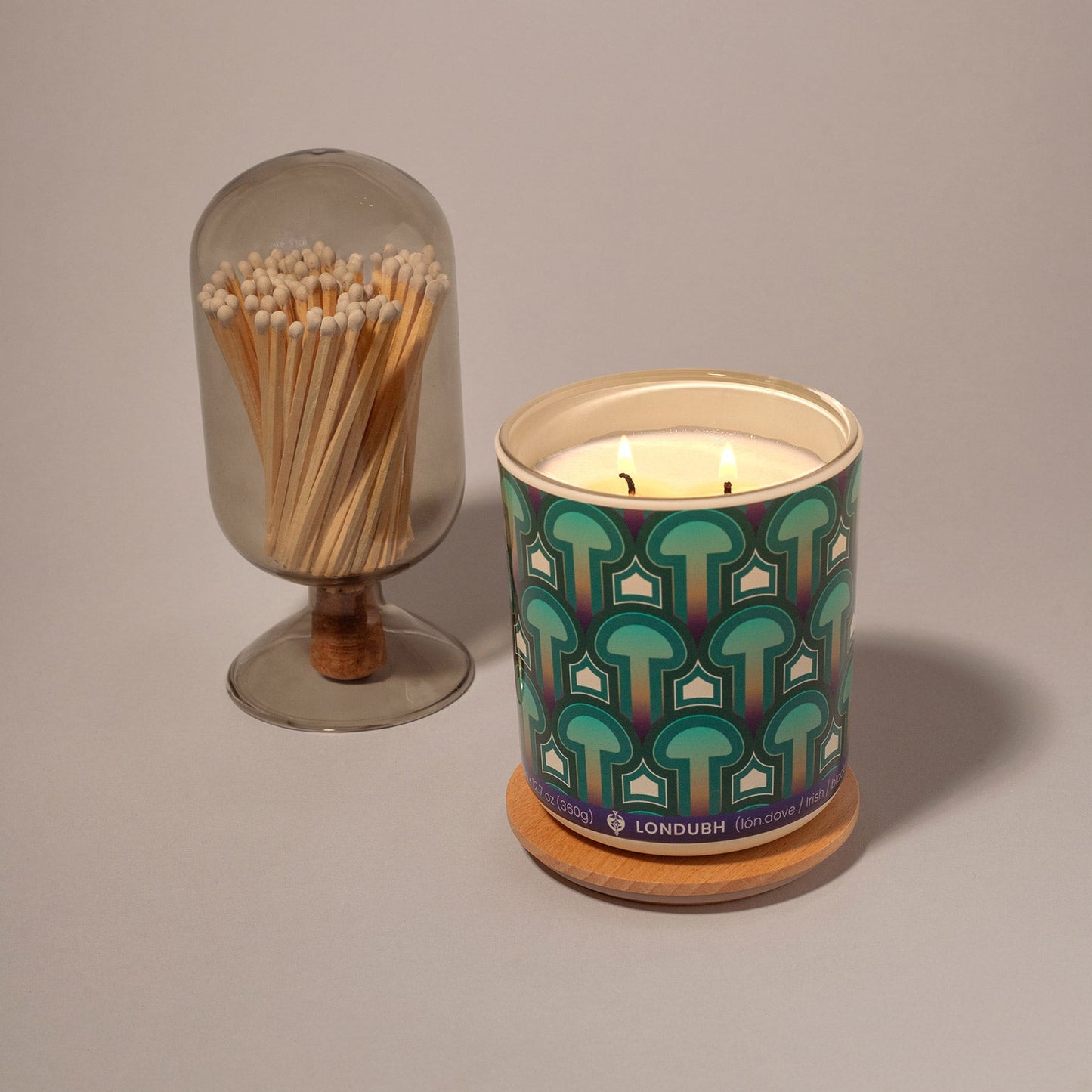A lit two-wick Enlightenment Candle in the 'Psilocybe' pattern, next to a glass cloche of matches.