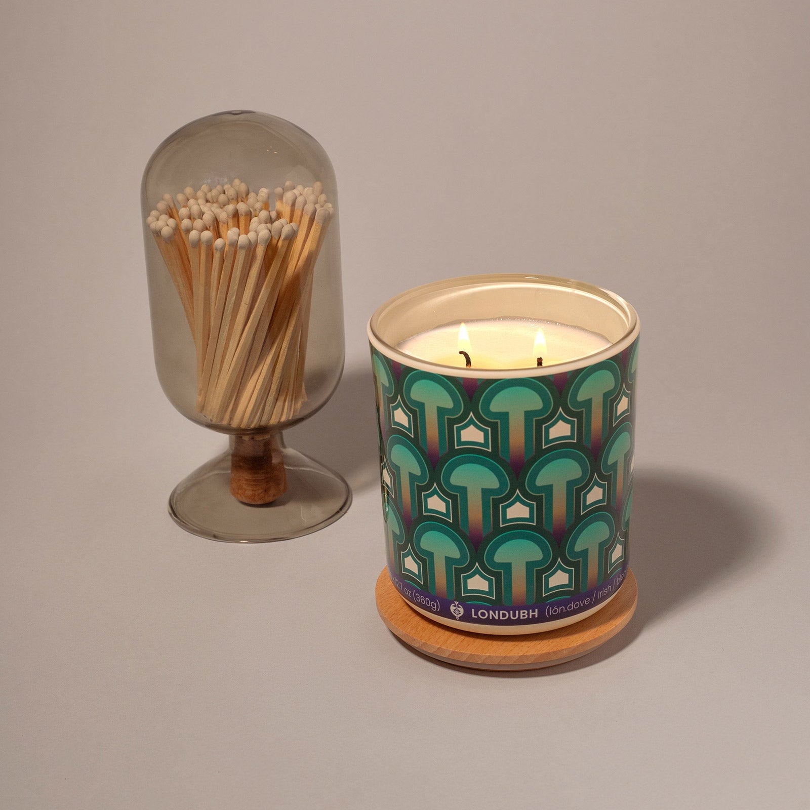 A lit two-wick Enlightenment Candle in the 'Psilocybe' pattern, next to a glass cloche of matches.