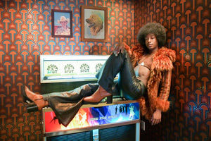 Black female moderl sitting on juke box in Desert 5 Spot at the tommy Hotel in Hollywood. Londubh Studio's enlightenment wallpaper in Panaeolous is on two walls wrapping a corner. Enlightenment Panaeolus is a shimmering repeat geometric pattern featuring mushrooms and the portals of discovery they inspire, in orange, red, pink, blue, colors inspired by David Bowie's Aladdin Sane album cover, all outlined in gold and printed on gold mylar