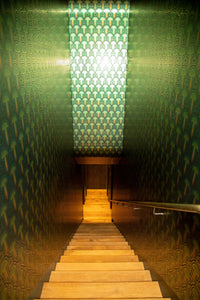 Installation of Enlightenment Psilocybe wallpaper on narrow staircase in Nicole Hollis designed private maximalist house. Enlightenment Psilocybe is a shimmering repeat geometric pattern featuring mushrooms and the portals of discovery they inspire, in blues, blue greens, a teal to gold sunset inspired ombre, all outlined in gold and printed on gold mylar