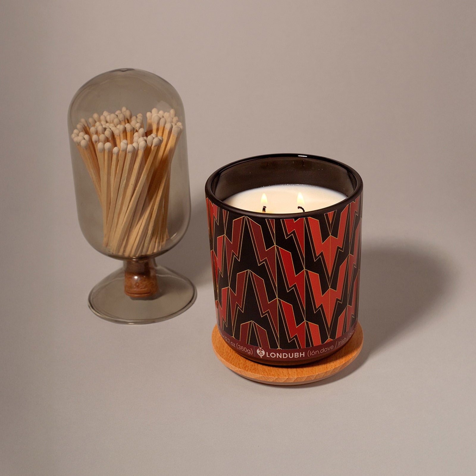 The lit Greased Lightning Candle sits on a cork coaster beside a glass cloche filled with long wooden matches.