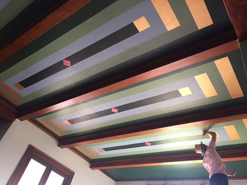 A person applies finishing touches to a hand-painted geometric ceiling with concentric rectangles.