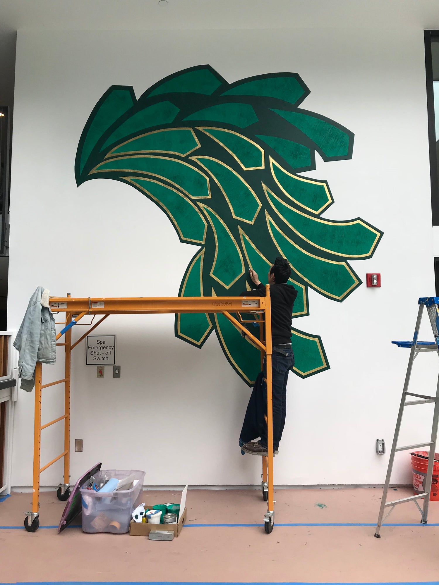 An artist on scaffolding carefully applies gold leaf detail to a large green abstract mural.