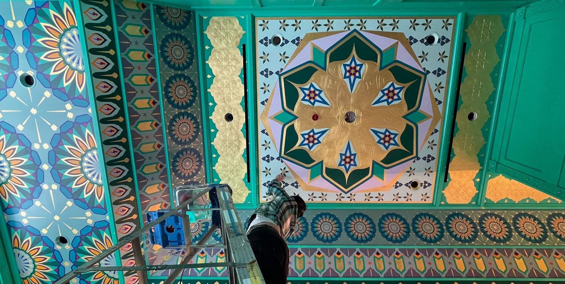 Founder Brynn Gelbard on a ladder hand-painting a vibrant geometric ceiling mural.