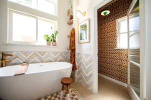 Wallpaper installation of custom grasscloth Neowise wallcovering installed on four walls and ceiling of small powder room shown with the door open from the larger modern bathroom angle. is a maximalist retro repeat pattern wallpaper inspired by a comet that flew over Newgrange in Ireland and looks like a feather motif but is inspired by grain, and is 70s browns, creams, and gold, and printed on gold mylar.