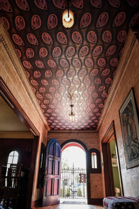 Peacock Nouveau aubergine purple, pink, and gold wallpaper shown installed on ceiling of house foyer and staircase against stone walls. Peacock Nouveau Aubergine is a Celtic and Art Nouveau inspired maximalist repeat pattern wallpaper featuring peacocks and Celtic knot work in pinks, purples, all outlined in gold and digitally printed on gold mylar.