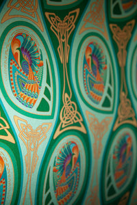 Peacock Nouveau Green Garden is a shimmering Celtic and Art Nouveau inspired maximalist repeat pattern wallpaper featuring peacocks and Celtic knot work in various greens, orange, blue, yellow, blues, purple, on a light green background, digitally printed on silver mylar - shown at an angle to highlight the shimmer