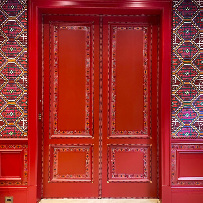 Ornate red double doors with intricate geometric borders next to a wall with a complex pattern.