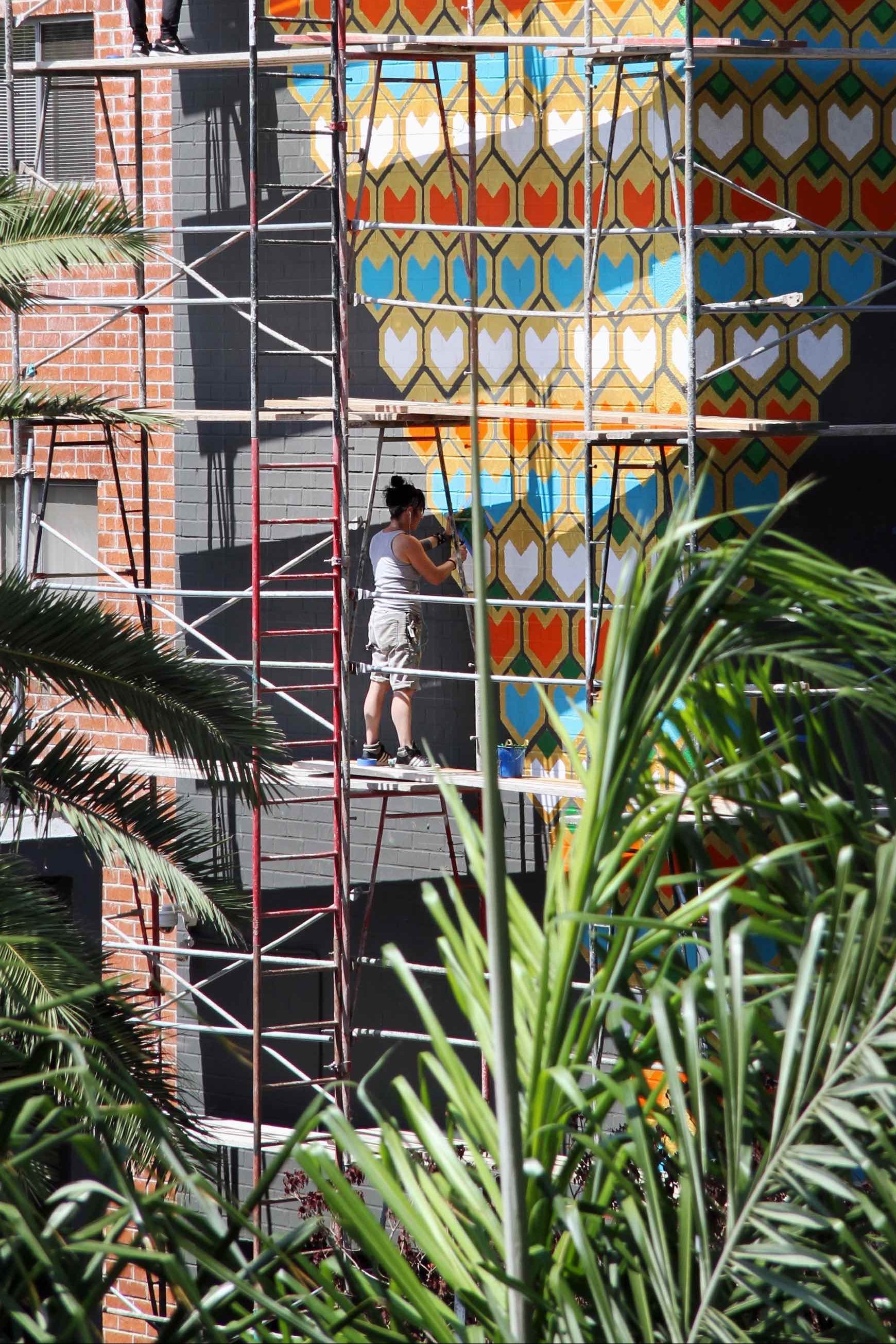 Artists on scaffolding paint a large, colorful geometric mural on a building facade.