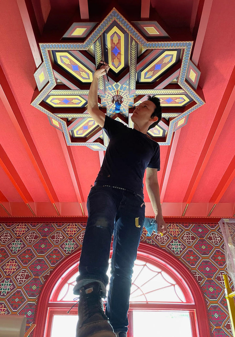 Lisa Donohoe of Londubh Studio standing on scaffolding painting a large ornate ceiling motif in tones of gold, black, red, white, and blue.
