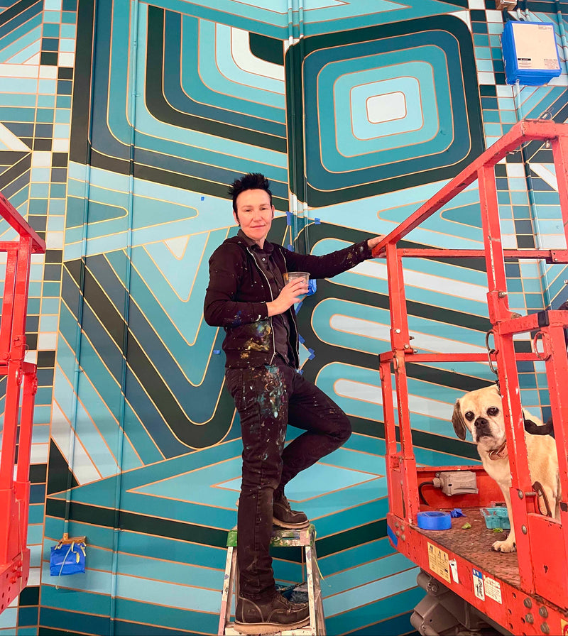 Founder Lisa Donohoe on a lift painting a large-scale geometric wall mural in blue and gold.