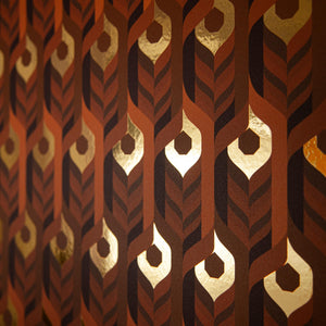 Neowise Radical is a maximalist retro repeat pattern wallpaper inspired by a comet that flew over Newgrange in Ireland and looks like a feather motif but is inspired by grain, and is 70s browns oranges, and gold, and printed on gold mylar - shown here at an angle with the light bouncing off the gold