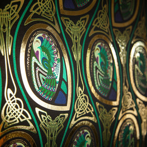 Peacock Nouveau Bejeweled is a Celtic and Art Nouveau inspired maximalist repeat pattern wallpaper featuring peacocks and Celtic knot work in gold, jewel tones, blues, greens, on a black background, outlined in gold and digitally printed on gold mylar - shown at an angle with the light bouncing off the gold