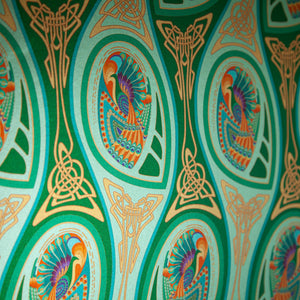 Peacock Nouveau Green Garden is a shimmering Celtic and Art Nouveau inspired maximalist repeat pattern wallpaper featuring peacocks and Celtic knot work in various greens, orange, blue, yellow, blues, purple, on a light green background, digitally printed on silver mylar  - shown at an angle to highlight the shimmer