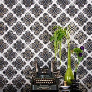 Blaze Inhale, a repeating geometric pattern featuring four black and white cannabis and marijuana leaves thinly framed in copper brown on a white background digitally printed on gold mylar shown illuminated in light bouncing off the shimmering gold trim outlining the pattern - shown in vignette with an Oliver #9 Typewriter and flowers in glass vases.