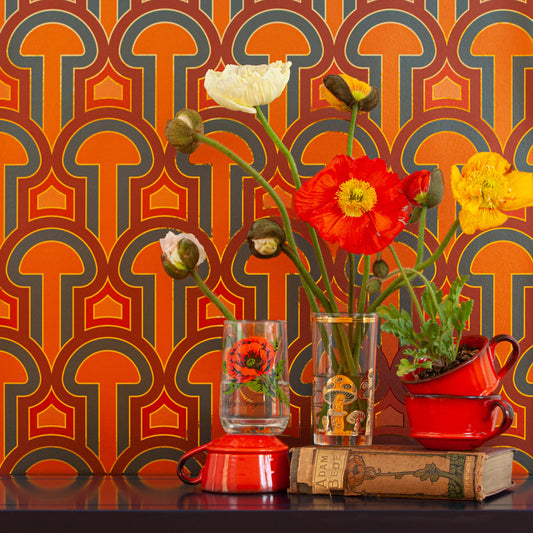 Enlightenment Panaeolus is a shimmering repeat geometric pattern featuring mushrooms and the portals of discovery they inspire, in orange, red, pink, blue, colors inspired by David Bowie's Aladdin Sane album cover, printed on gold mylar - shown here in a vignette with flowers in glasses and old books