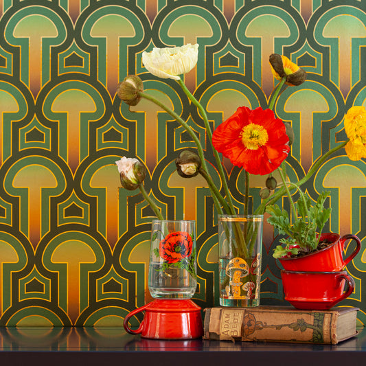 Enlightenment Psilocybe is a shimmering repeat geometric pattern featuring mushrooms and the portals of discovery they inspire, in blues, blue greens, a teal to gold sunset inspired ombre, all outlined in gold and printed on gold mylar - shown here in a vignette with flowers in glasses and old books