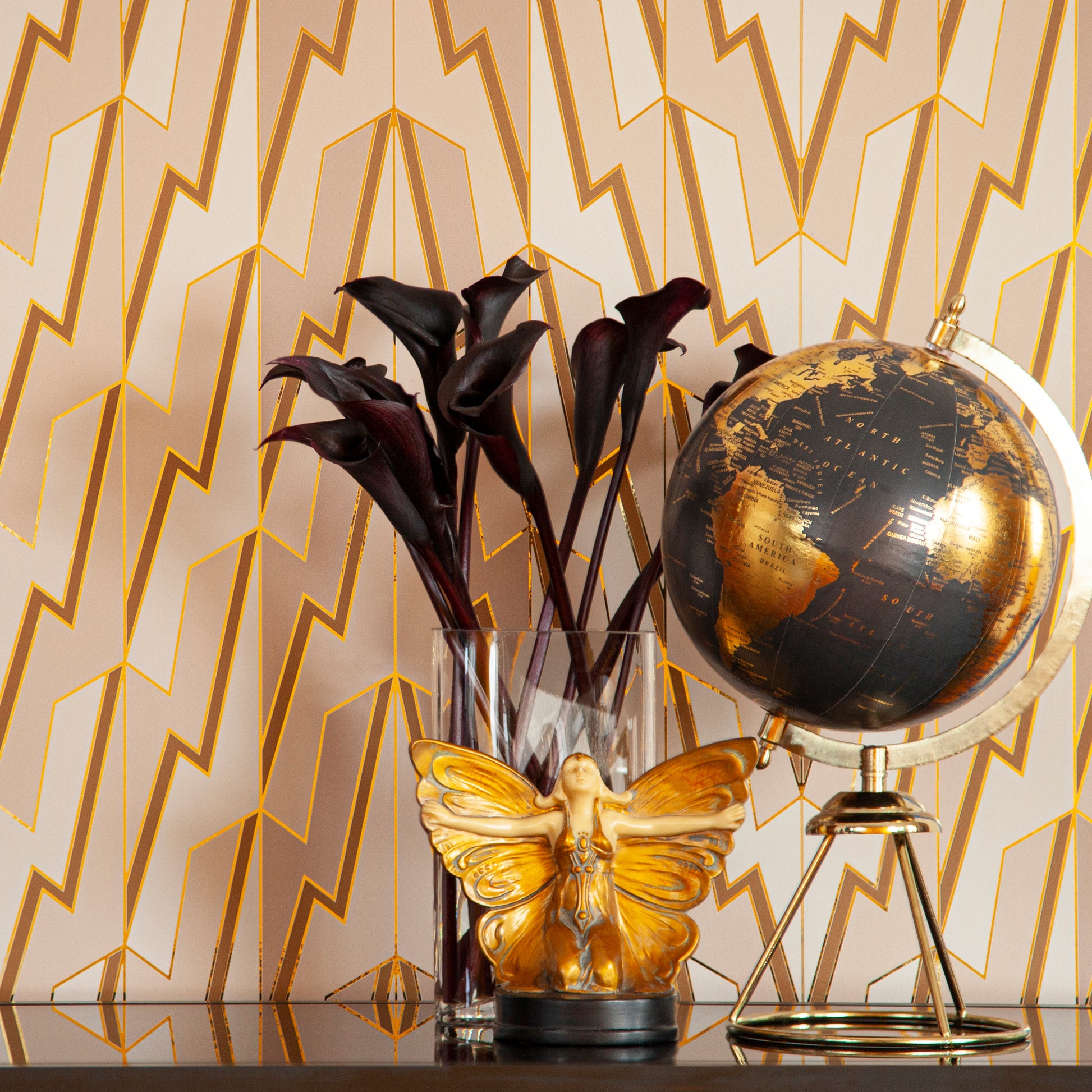 Greased Lightening Cream Dreams is a repeat art deco inspired gothamesque geometric pattern showcasing the lightning bolt, in neutral warm whites, beiges, and tans, outlined in gold and printed on gold mylar - shown here in a vignette with a little butterfly statue, black flowers and a vintage globe
