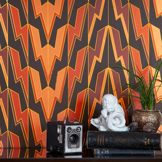 Greased Lightening Power of Love is an  art deco inspired gothamesque geometric repeat pattern showcasing the lightning bolt, in black, reds, pinks, outlined in gold and printed on gold mylar - shown in a vignette with an antique book, a monkey statue, a vintage box camera and a plant