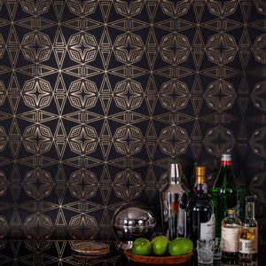 Integratronic Night is a mid-century retro repeat geometric pattern featuring dark green and gray motifs on a black background all outlined in gold and digitally printed on gold mylar  - shown in a vignette with a bar set up with bottles, limes, and shot glasses