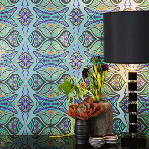 Ulu Garden is a graphic repeat pattern featuring various gold outlined organic motifs geometrically woven together in various vibrant blues, greens, and floral colors orange, purples, outlined in gold, digitally printed on gold mylar, shown in a vignette with flowers, a black lamp, and pottery bowls.