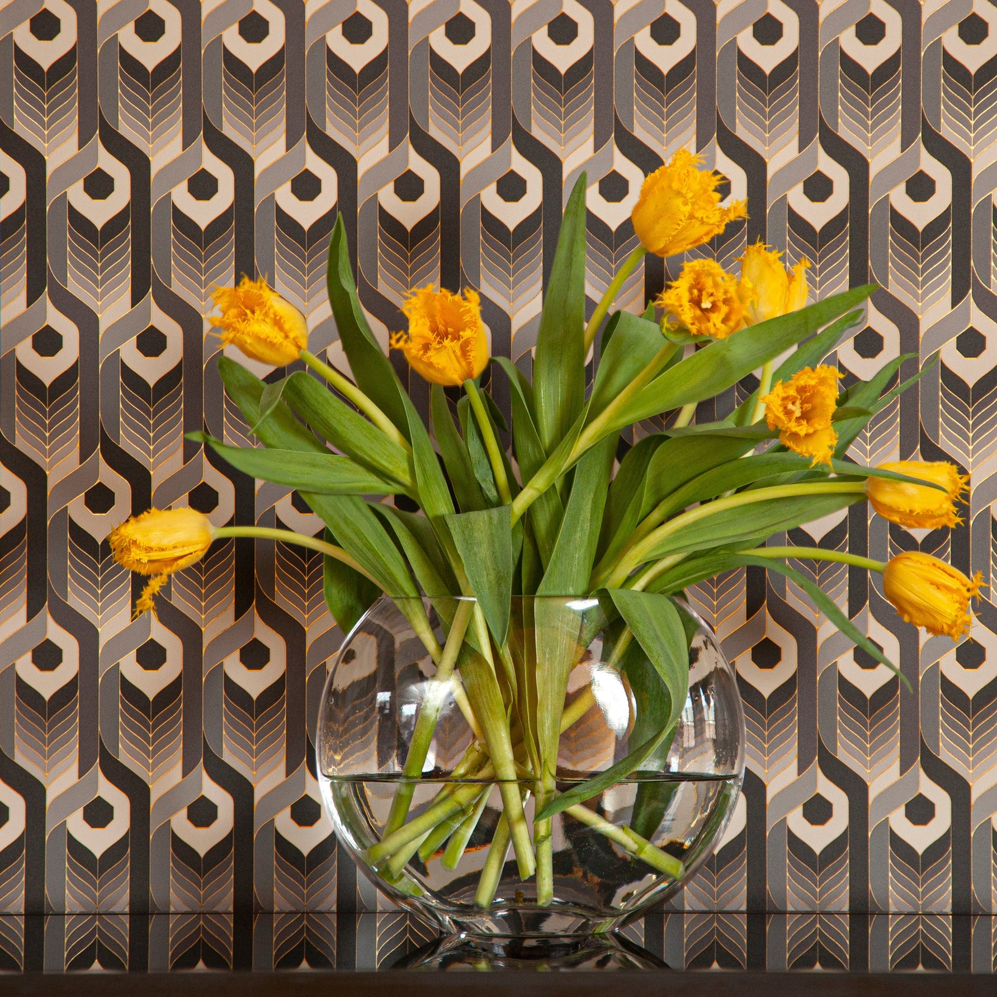 Neowise Classical is a repeat pattern wallpaper inspired by Neowise, a comet that flew over Newgrange in Ireland.  Neowise Classical looks like a feather motif but is inspired by grain, and is black, an ombre of gray tones, white, outlined in gold, and printed on gold mylar - shown here with a glass bowl of yellow flowers