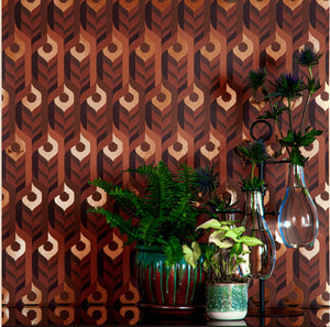 Neowise Radical is a maximalist retro repeat pattern wallpaper inspired by a comet that flew over Newgrange in Ireland and looks like a feather motif but is inspired by grain, and is 70s browns oranges, and gold, and printed on gold mylar - shown in a vignette with plants