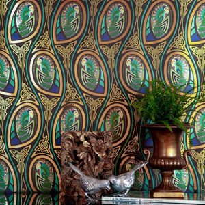 Peacock Nouveau Bejeweled is a Celtic and Art Nouveau inspired maximalist repeat pattern wallpaper featuring peacocks and Celtic knot work in gold, jewel tones, blues, greens, on a black background, outlined in gold and digitally printed on gold mylar  - shown in a vignette with plants, a block of gnarled wood, a book and a metal sculpture of 2 birds 