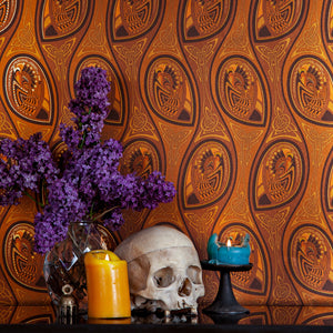 Peacock Nouveau Jackie Brown is a shimmering Celtic and Art Nouveau inspired maximalist repeat pattern wallpaper featuring peacocks and Celtic knot work in 70s retro browns, golds, and oranges, digitally printed on gold mylar - shown in a vignette with candles, purple flowers, and a skull